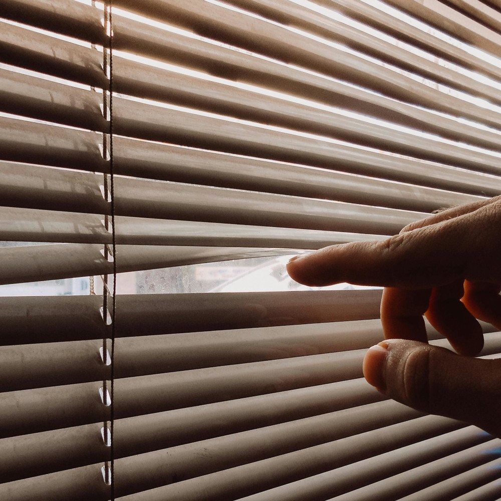 A close-up of a hand using two fingers to pull apart horizontal window blinds to peek outside.