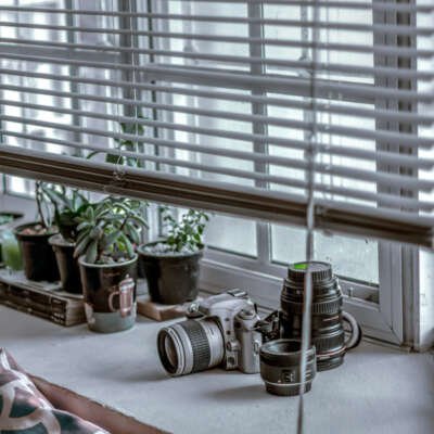A Nikon DSLR camera and multiple lenses sitting on a white windowsill behind horizontal blinds, with small potted plants in the background.